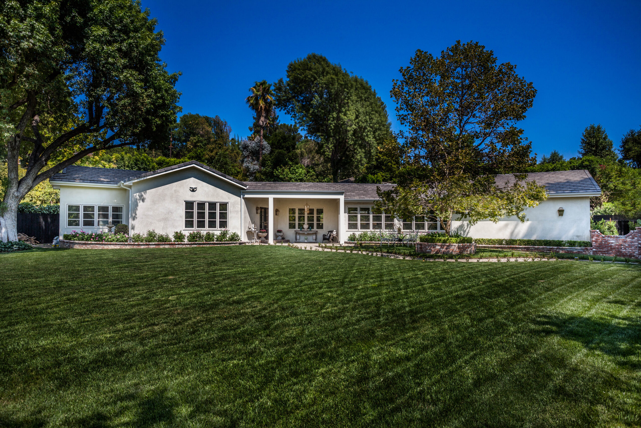 Home Building A single-story white house with a well-manicured lawn and surrounded by trees under a clear blue sky.