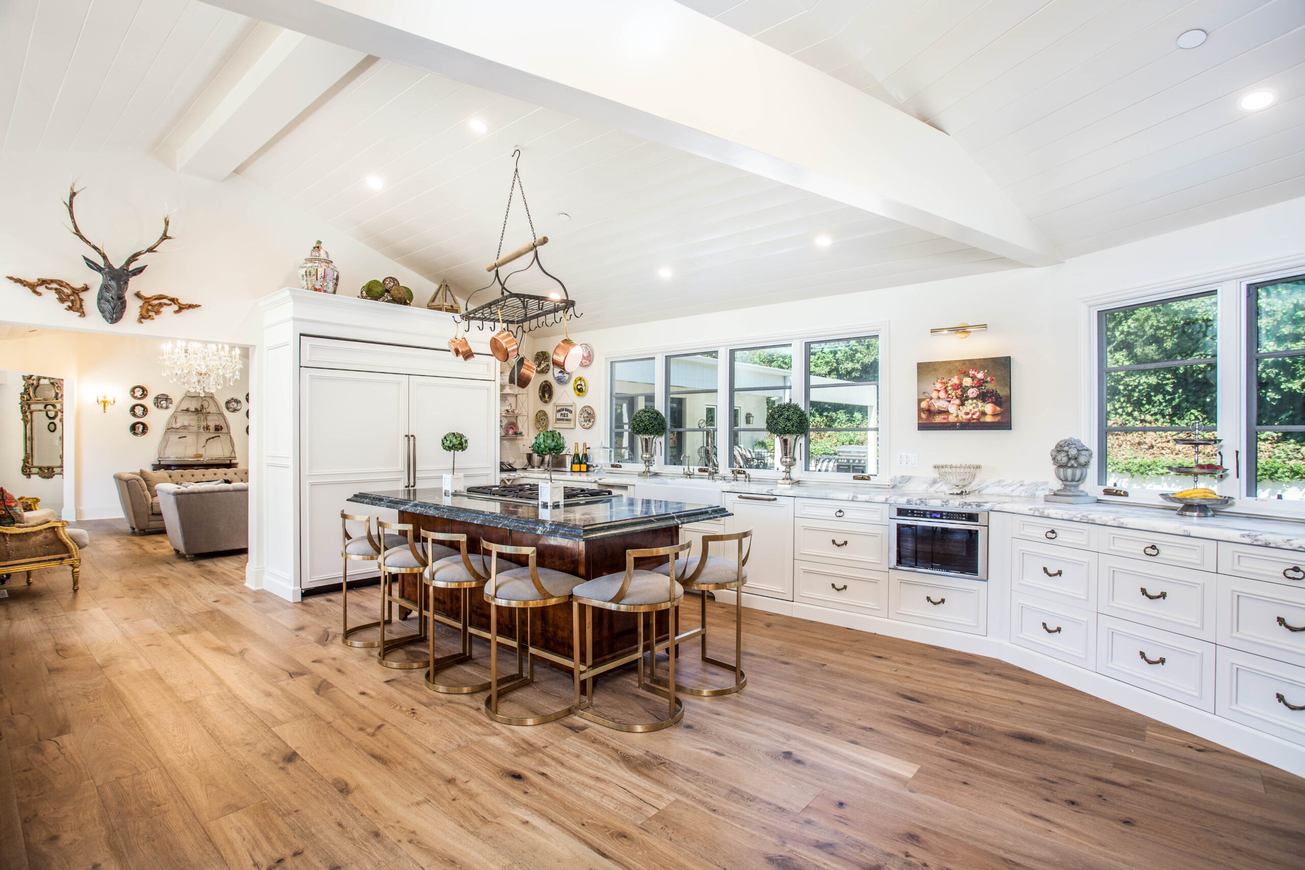 Bright kitchen with white cabinets, a large island, bar stools, wood floor, and wall decor; windows overlook a garden.