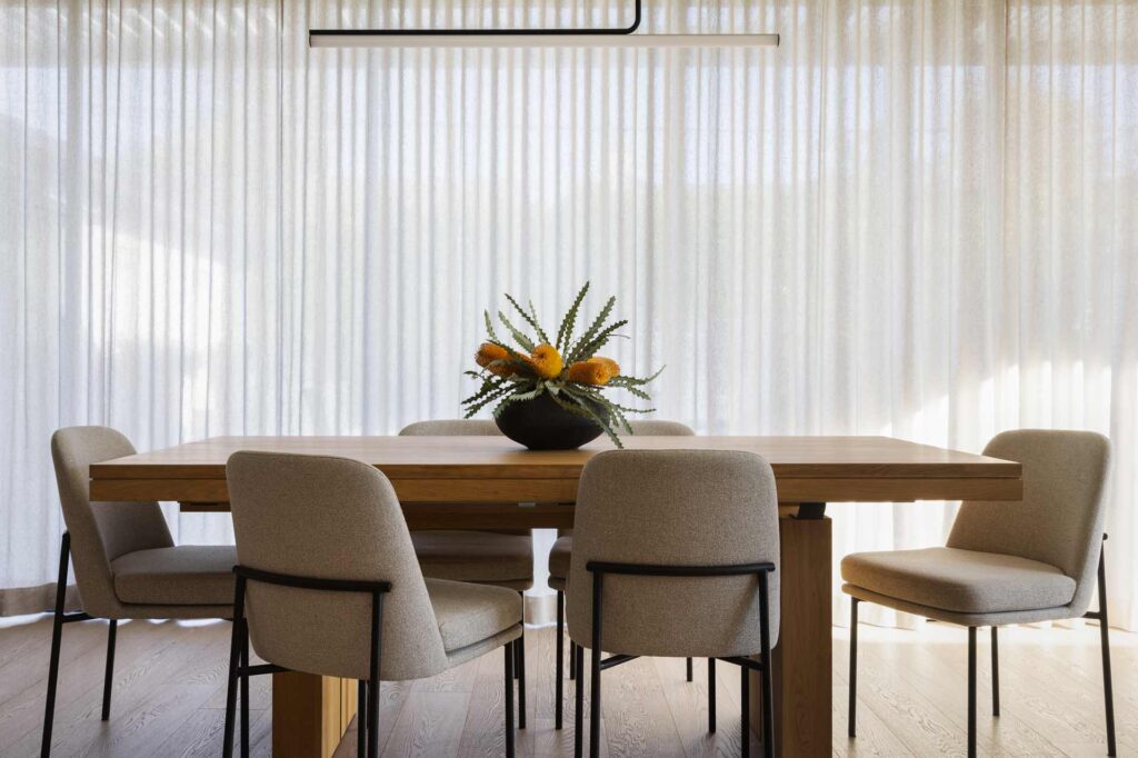 Dining room with a wooden table, six beige chairs, and a vase with yellow flowers. Sheer curtains in the background offer inspiration for kitchen remodeling tips that blend elegance and warmth.