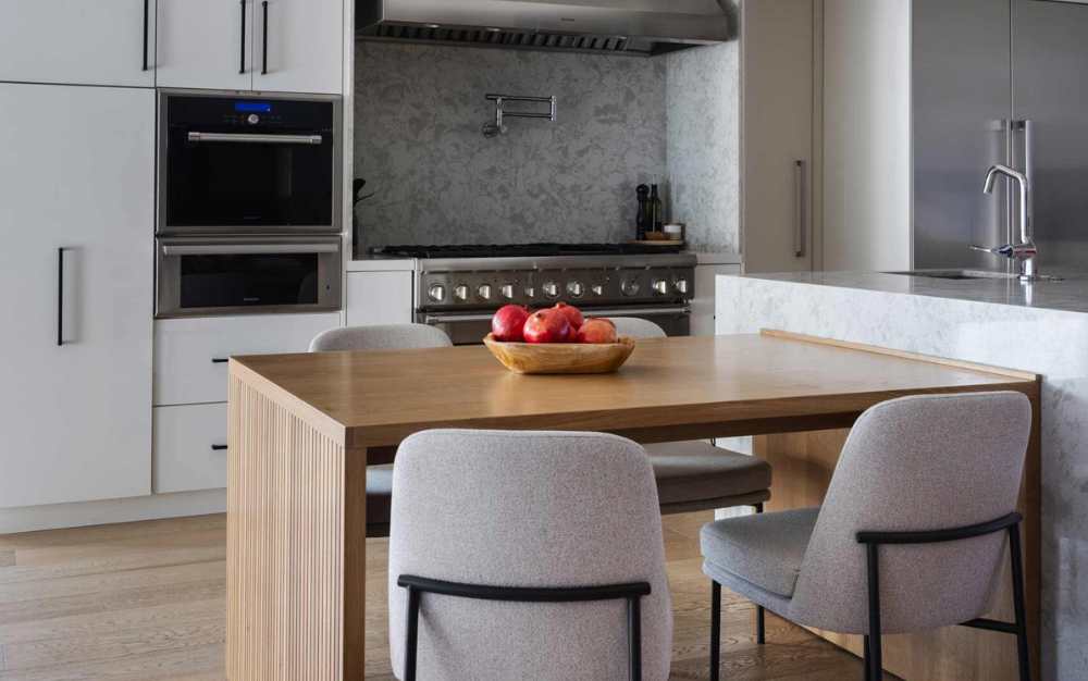 Modern kitchen remodel featuring a wooden table, gray chairs, a bowl of apples, marble backsplash, and sleek stainless steel appliances.