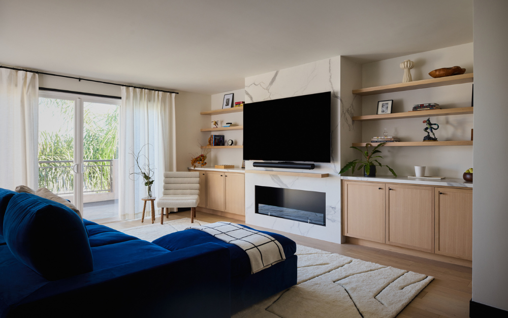 Building Contractors This modern living room, crafted with the expertise of building contractors, features a blue couch, a large TV, and a cozy fireplace. Light wood cabinets and shelves complement the space, while white curtains and a rug add a touch of elegance.