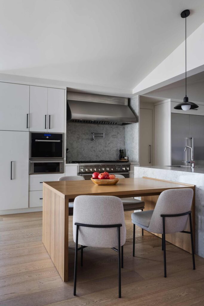 Kitchen Remodel Project Modern kitchen with a wooden table, two chairs, bowl of apples, white cabinets, stove, and overhead light. This stylish space reflects a recent kitchen remodel with its sleek design and fresh ambiance.