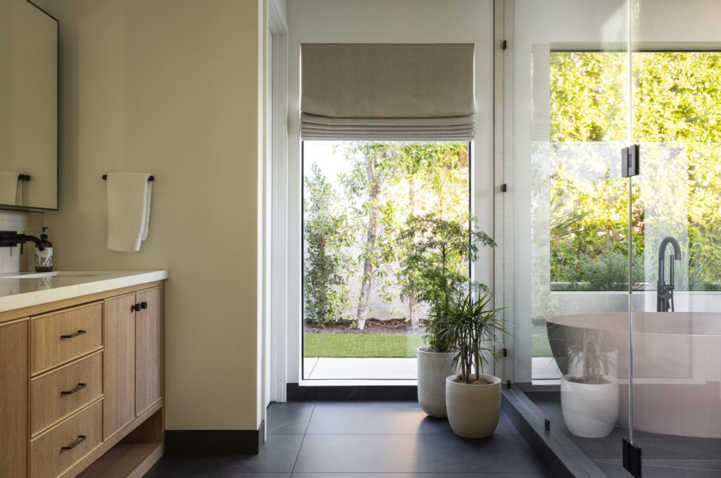 Modern bathroom with wood vanity, potted plants, and glass shower, brightened by a large window with garden view—a perfect example of expert bathroom remodeling services.