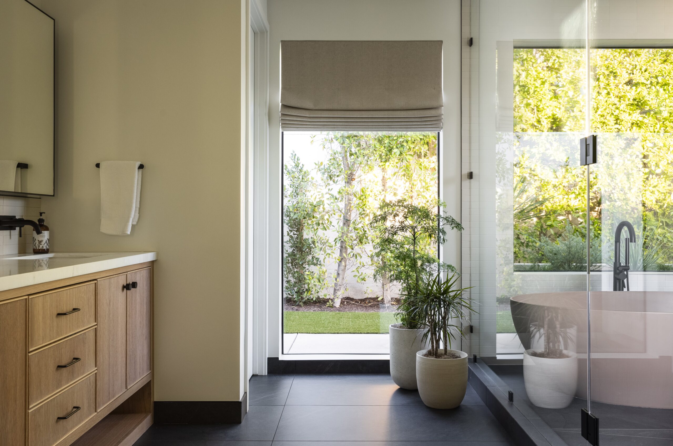 Modern bathroom with wood vanity, potted plants, and glass shower, brightened by a large window with garden view—a perfect example of expert bathroom remodeling services.