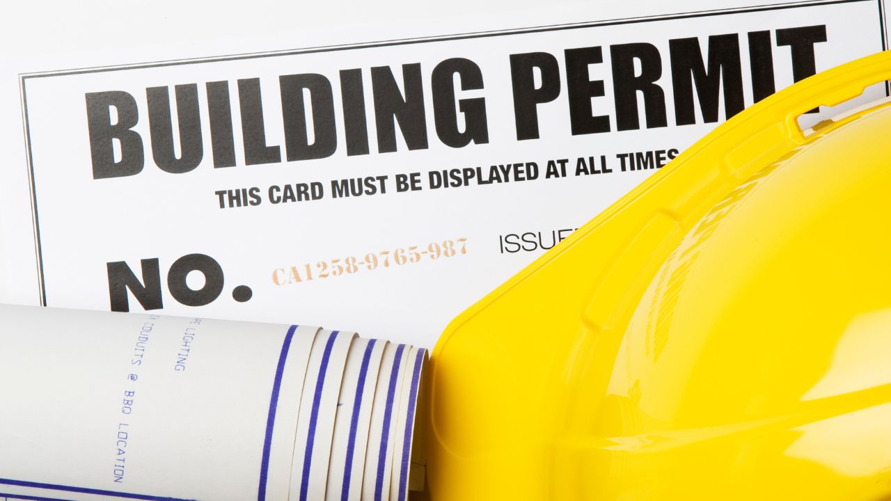 A building permit, construction blueprint, and yellow hard hat arranged together, representing general contractors at work.