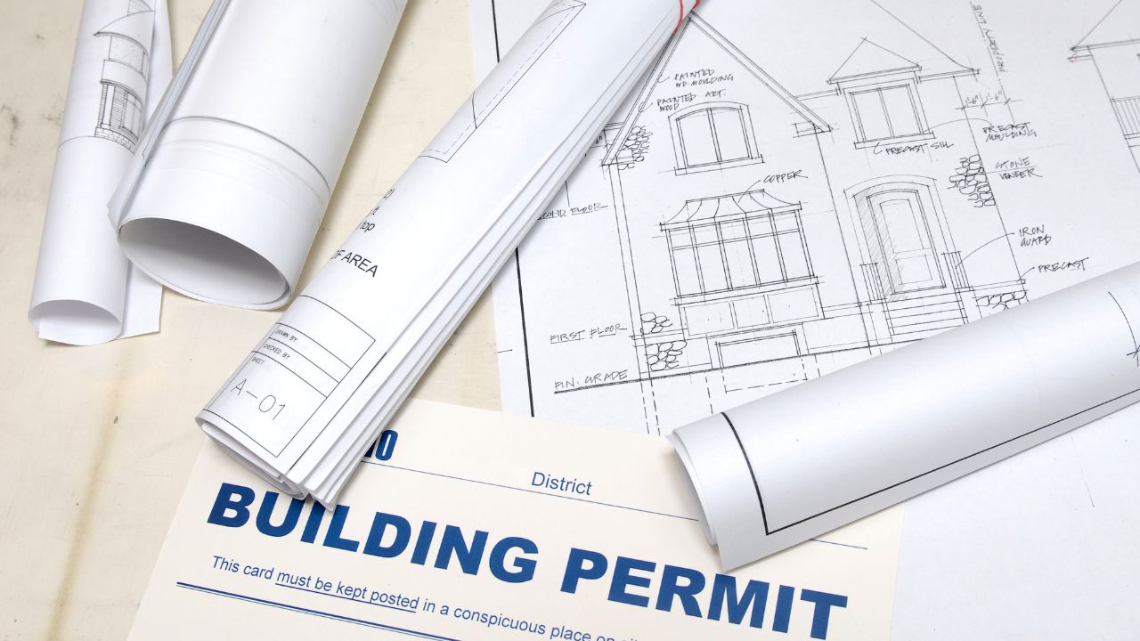 Rolled blueprints and a building permit lying on top of an architectural house drawing, illustrating the careful planning a custom home builder brings to every project.