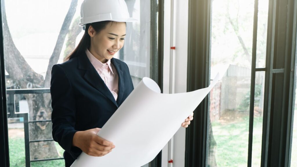 A woman in a suit and hard hat smiles as she reviews a large blueprint near a window, embodying the confidence and expertise of a custom home builder.