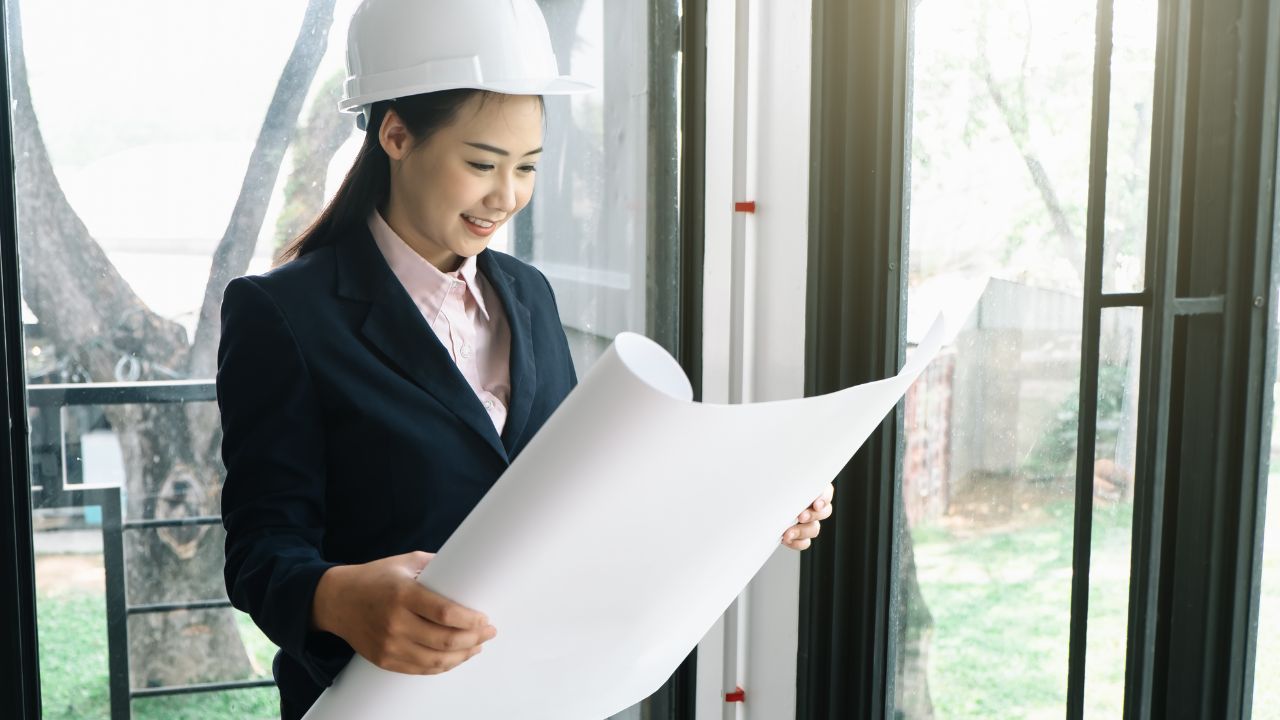 A woman in a suit and hard hat smiles as she reviews a large blueprint near a window, embodying the confidence and expertise of a custom home builder.