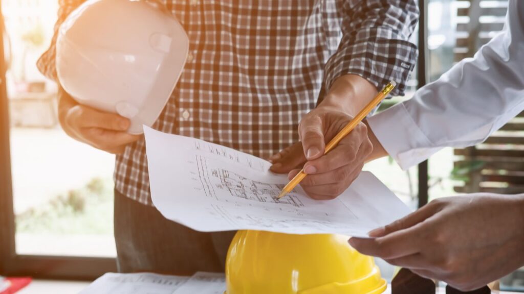 Two people reviewing construction plans; one holds a hard hat, while the other, likely from a team of custom home builders, points at detailed drawings with a pencil.