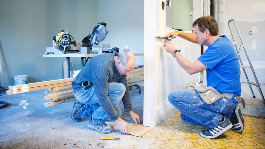 Two men with a General Contractor (GC) license install a door frame in a room under renovation, surrounded by tools and wood pieces.
