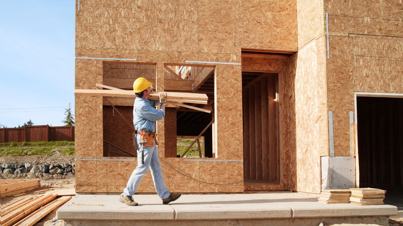 Construction worker in a hard hat carries wooden planks in front of a partially built green custom home in Los Angeles.